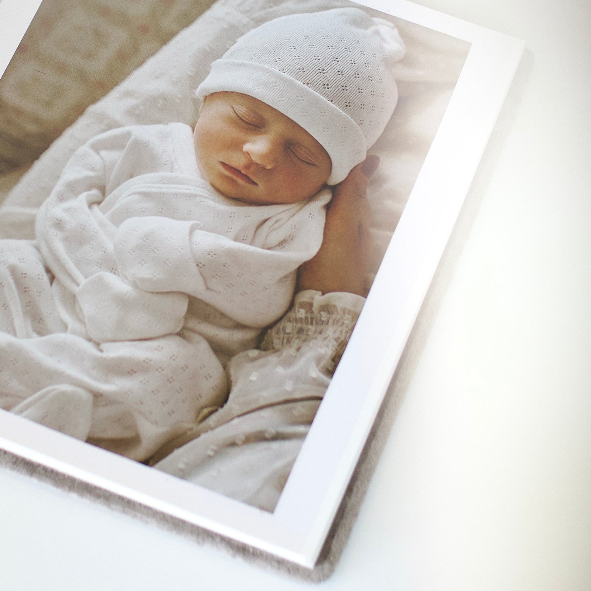 Newborn baby wrapped in white with a matching hat, lying on a soft surface. Preview of how a photo may appear inside the album.
