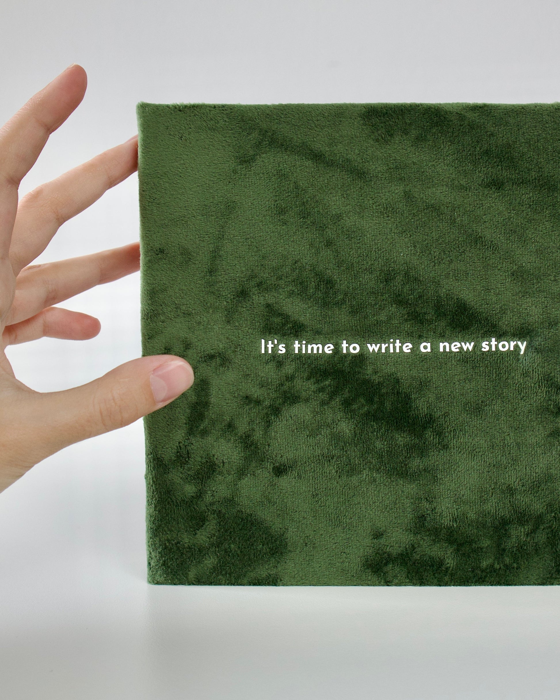 Green notebook with 'It's time to write a new story' text, held by a hand on a white background