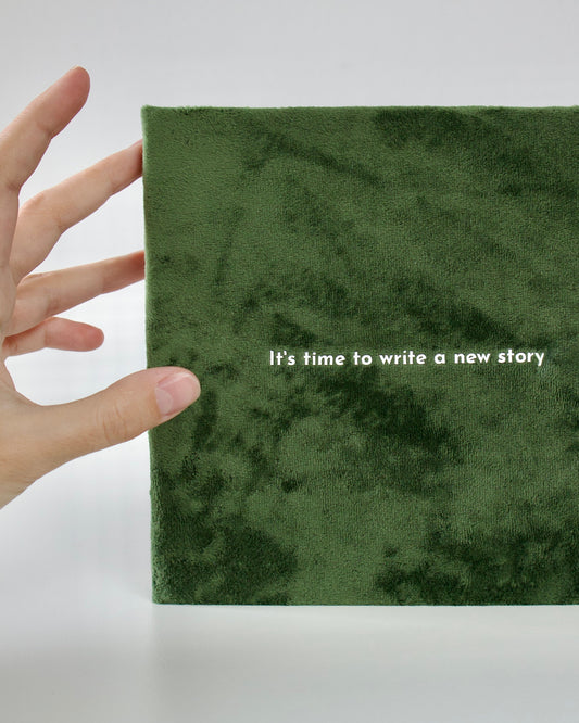Green notebook with 'It's time to write a new story' text, held by a hand on a white background